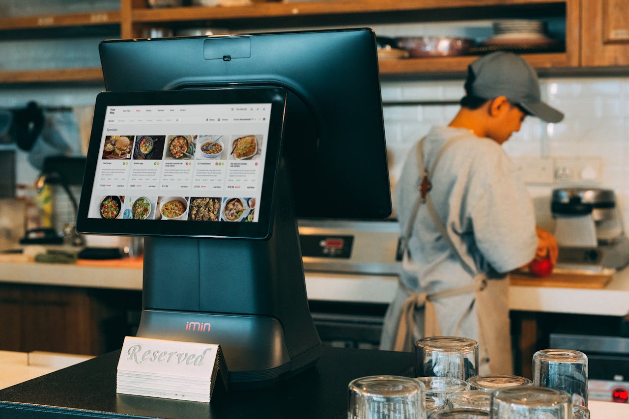 about-us Digital ordering system in a restaurant kitchen with a touchscreen display and a chef at work.
