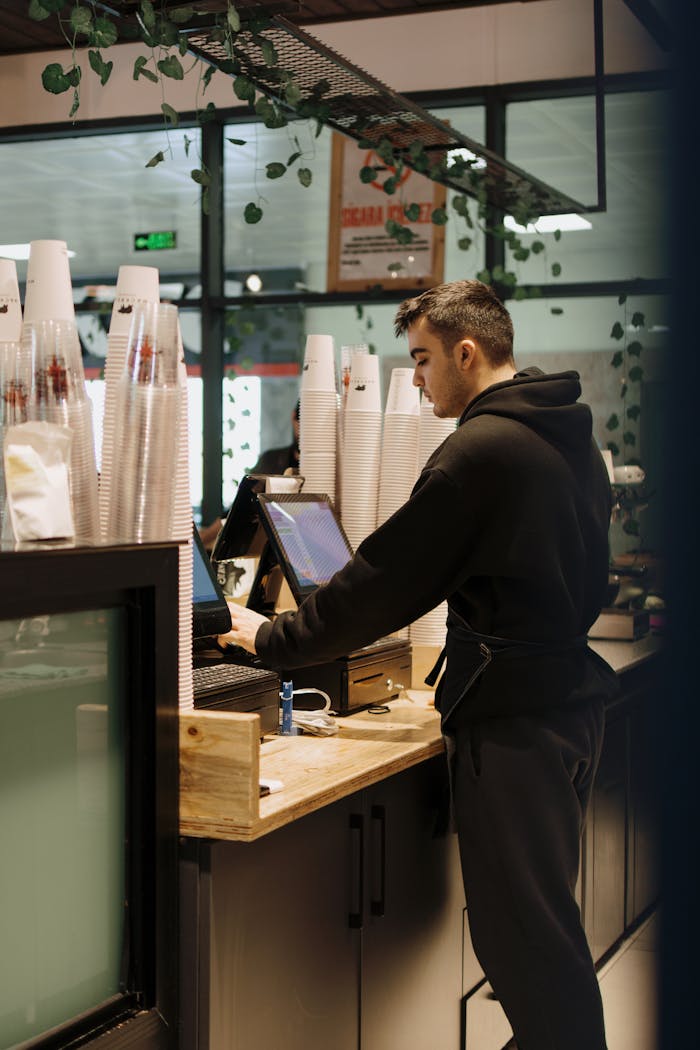 services-04 A barista in a black hoodie working at the register in a contemporary café with coffee cups stacked nearby.