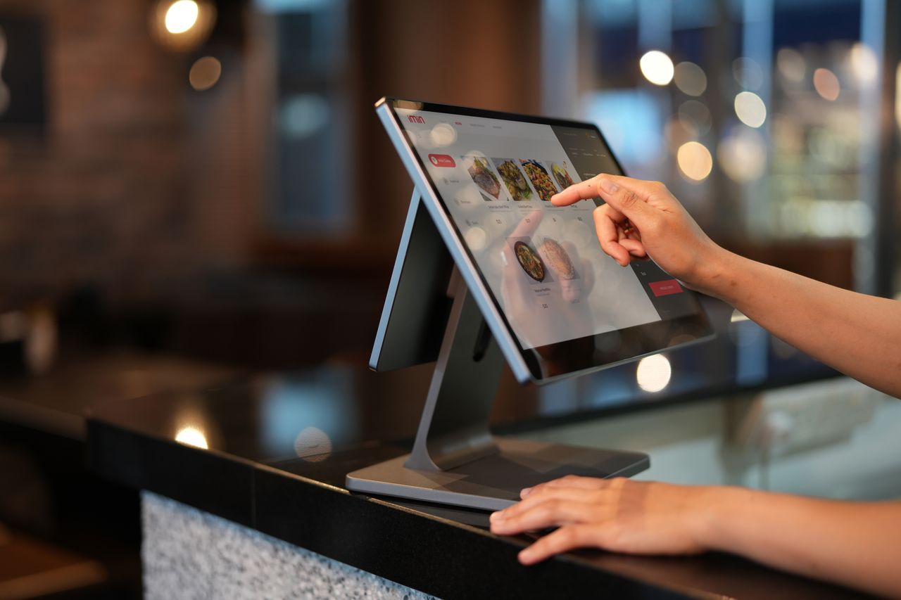 services-01 Close-up of a hand interacting with a touchscreen POS system in a restaurant setting.