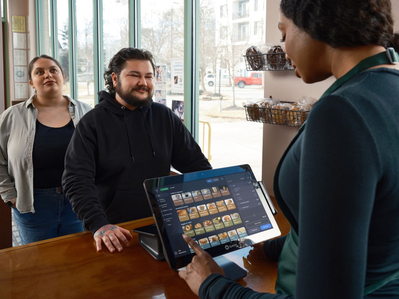 about-us-01 Barista using POS system to serve two customers at a café counter, showcasing modern restaurant technology.